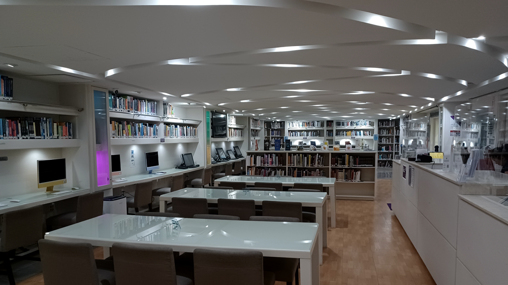 English: Modern university library with study tables, desktop computers, and bookshelves filled with books. Español: Biblioteca universitaria moderna con mesas de estudio, computadoras de escritorio y estanterías llenas de libros.