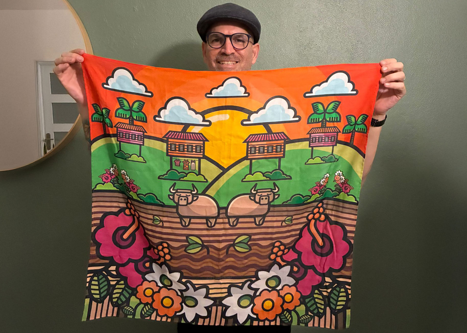 El profesor Miguel Morales sostiene frente a sí un colorido pañuelo de seda con una ilustración inspirada en el paisaje rural puertorriqueño. El diseño muestra casas de campo, palmas, dos bueyes sobre terreno arado, flores tropicales y un cielo anaranjado con nubes. Morales sonríe mientras posa frente a una pared verde.
