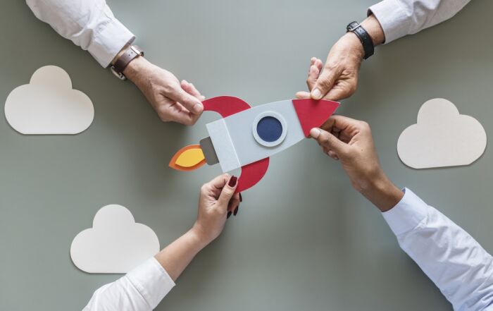 Español: Manos de varias personas colaborando para armar un cohete de papel sobre una superficie verde, con figuras de nubes alrededor, representando trabajo en equipo, creatividad y el lanzamiento de una idea o proyecto. English: Hands of several people collaborating to assemble a paper rocket on a green surface, with cloud shapes around it, representing teamwork, creativity, and the launch of an idea or project.