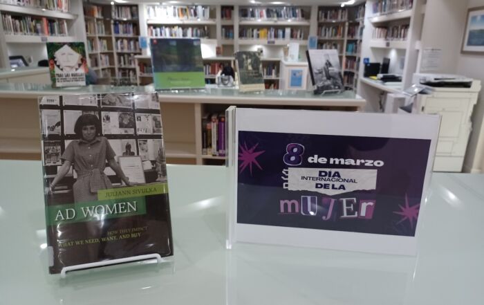 Mesa de exhibición en una biblioteca con el libro Ad Women de Juliann Sivulka y un letrero que dice “8 de marzo – Día Internacional de la Mujer”, con estanterías de libros al fondo.