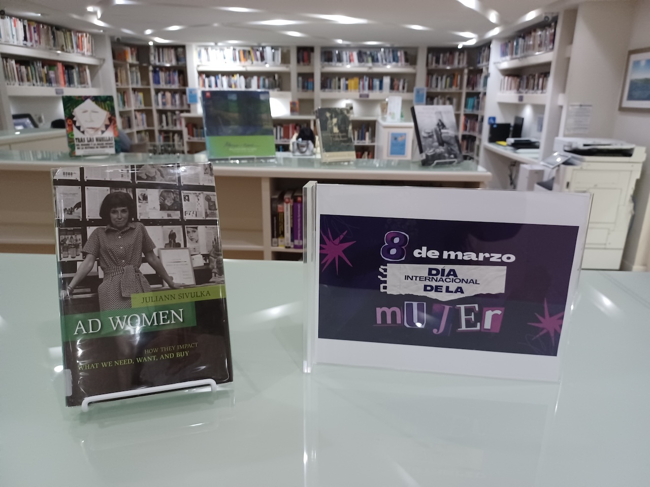 Mesa de exhibición en una biblioteca con el libro Ad Women de Juliann Sivulka y un letrero que dice “8 de marzo – Día Internacional de la Mujer”, con estanterías de libros al fondo.