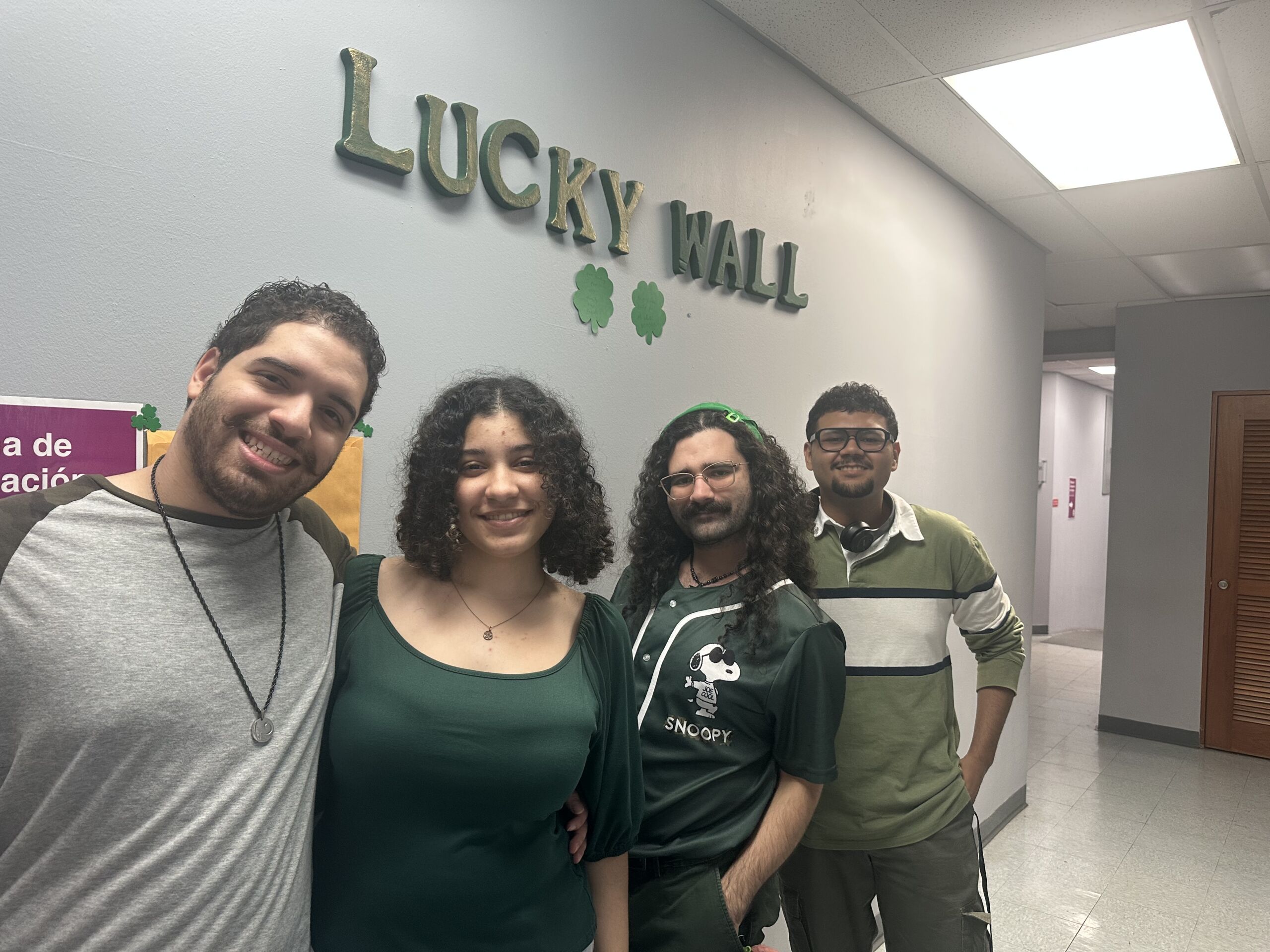 Cuatro personas vestidas de verde posan en un pasillo frente a una pared con el rótulo “Lucky Wall” y decoraciones de tréboles.