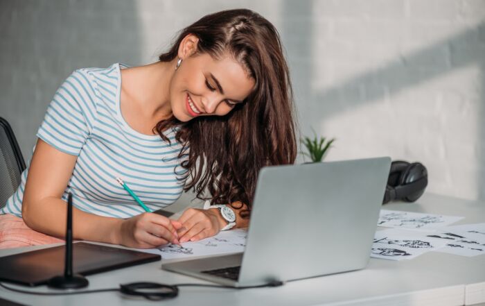 English (Alt Text): Young woman smiling while sketching drawings on paper at a desk with a laptop and graphic tablet, working in a creative design workspace. Español (Texto Alternativo): Joven sonriente dibujando bocetos en papel en un escritorio con una laptop y una tableta gráfica, trabajando en un espacio creativo de diseño.