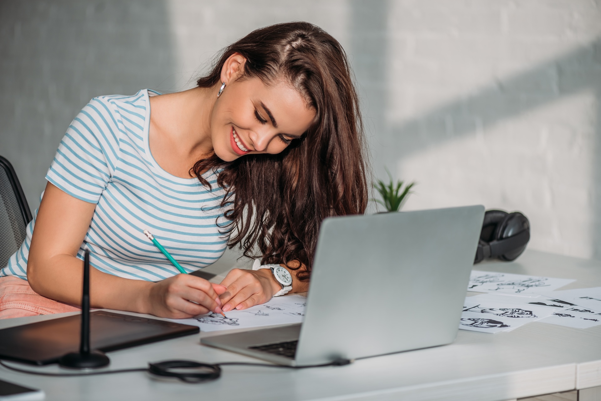 English (Alt Text): Young woman smiling while sketching drawings on paper at a desk with a laptop and graphic tablet, working in a creative design workspace. Español (Texto Alternativo): Joven sonriente dibujando bocetos en papel en un escritorio con una laptop y una tableta gráfica, trabajando en un espacio creativo de diseño.